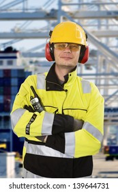 Portrait Of A Confident Docker, Wearing All Required Personal Protective Equipment, Posing In Front Of An Industrial Container Terminal And Harbor