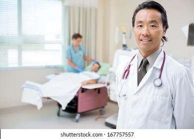 Portrait Of Confident Doctor In Hospital Room With Nurse Giving Dialysis To Patient In Background