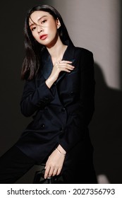 Portrait of confident elegant business woman wearing navy blue suit isolated on dark grey background. Young female model with black hair posing in fashion while sitting on stool looking at the camera.
