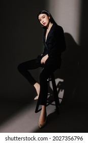 Portrait Of Confident Elegant Business Woman Wearing Navy Blue Suit Isolated On Dark Grey Background. Young Female Model With Black Hair Posing In Fashion While Sitting On Stool Looking At The Camera.