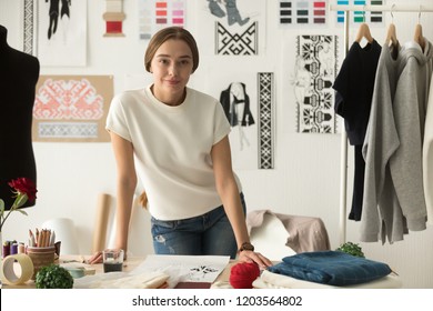 Portrait of confident fashion clothing designer standing at workplace leaning on desktop, talented young dressmaker, tailor shop owner posing in workshop, sewing shop, studio, looking at camera
