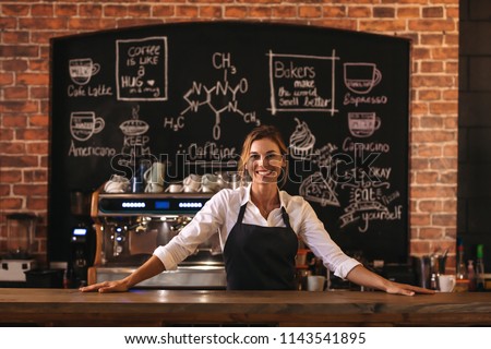 Portrait of confident female barista standing behind counter. Woman cafe owner in apron looking at camera and smiling.