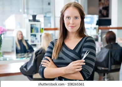 Portrait of a confident female hairdresser standing hands folded with people in background - Powered by Shutterstock