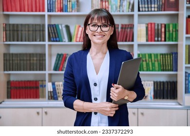 Portrait of confident female teacher holding laptop inside library