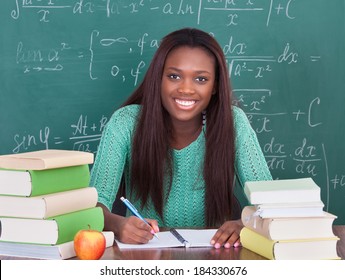 Portrait of confident female teacher writing in book at classroom desk