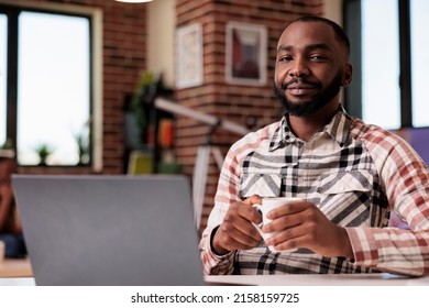 Portrait Of Confident Freelance Programmer Looking At Camera Taking A Break From Coding Holding A Cup Of Coffee. African American Student Relaxing At Home Watching Social Media Content On Laptop.