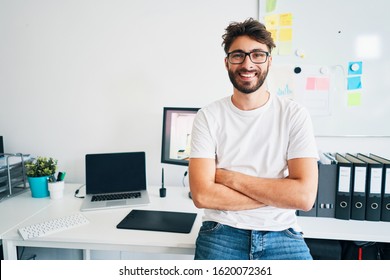 Portrait Of Confident Graphic Designer Leaning On Desk In Office With Arms Crossed