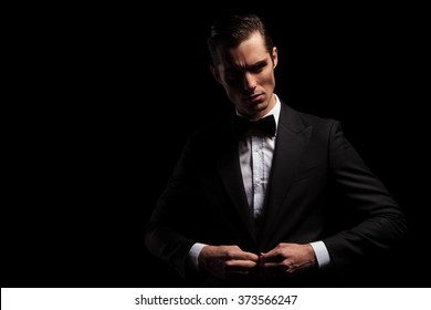 Portrait Of Confident Handsome Man In Black Suit With Bowtie Posing In Dark Studio Background While Closing His Jacket 