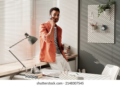 Portrait Of Confident Handsome Young Black Man In Stylish Outfit Standing With Takeout Coffee At Window And Chatting By Phone In Office