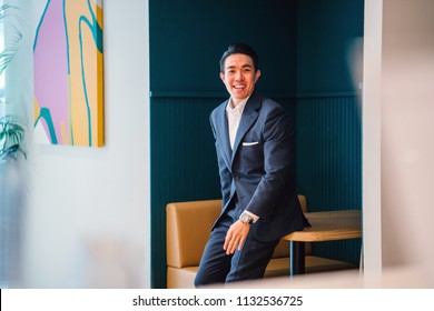 Portrait Of A Confident, Handsome Young Chinese Asian Professional Man Wearing A Suit And Leaning Against A Table In His Office. He Is Athletic And Muscular And Smiling As He Smiles For His Head Shot.