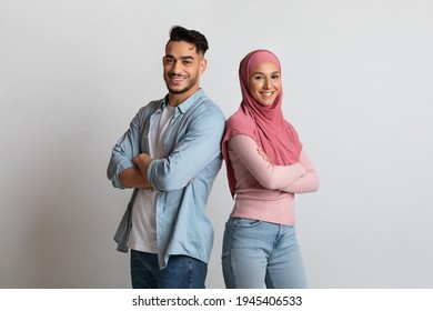 Portrait Of Confident Happy Islamic Couple Standing Back To Back With Folded Arms, Positive Arab Man And Muslim Woman In Hijab Posing Together On Grey Studio Background, Looking And Smiling At Camera