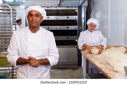 Portrait Of Confident Hispanic Bakery Owner In White Uniform Posing Against Busy Workers Background