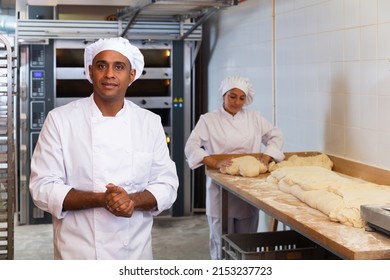 Portrait Of Confident Hispanic Bakery Owner In White Uniform Posing Against Busy Workers Background