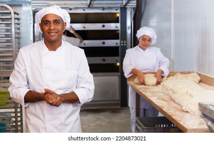 Portrait Of Confident Hispanic Bakery Owner In White Uniform Posing Against Busy Workers Background