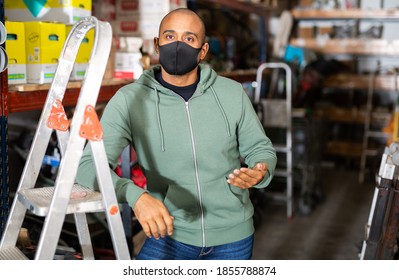 Portrait Of Confident Hispanic Man Owner Of Hardware Store In Protective Face Mask Posing Among Shelving With Goods. New Lifestyle In Concept Of Coronavirus Pandemic