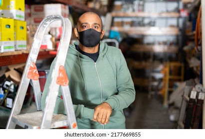 Portrait Of Confident Hispanic Man Owner Of Hardware Store In Protective Face Mask Posing Among Shelving With Goods. New Lifestyle In Concept Of Coronavirus Pandemic