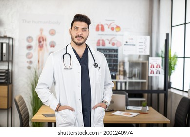 Portrait Of Confident Indian Doctor Keeping Hands In Pockets Of White Lab Coat While Posing At Hospital Room. Concept Of People, Health Care And Treatment.