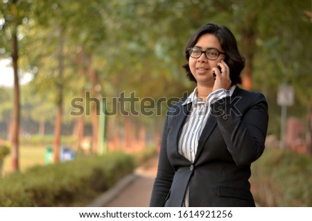 Portrait of a confident looking young Indian woman with short hair and spectacles speaking on a mobile phone in outdoor setting wearing a formal striped shirt and black blazer in a park