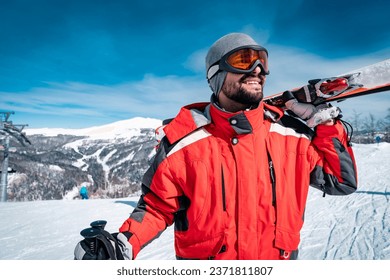 Portrait of confident male skier in red jacket standing on top of mountain while holding skies. Happy male enjoying ski holiday during the sunny winter day. - Powered by Shutterstock