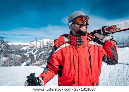 Portrait of confident male skier in red jacket standing on top of mountain while holding skies. Happy male enjoying ski holiday during the sunny winter day.