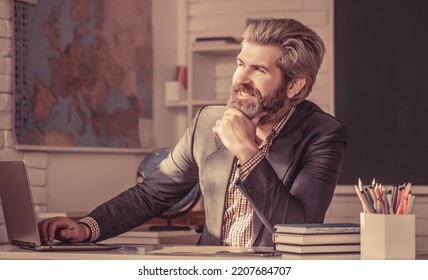 Portrait Of Confident Male Teacher In Classroom. Young Male Teacher With Laptop In Class Sits At The Table The Blackboard In The Classroom.