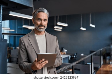 Portrait of confident mature business man executive standing in corporate office using digital tablet. Middle aged professional businessman manager wearing suit posing at work in modern space.