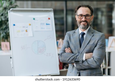 Portrait of a confident mature businessman standing with arms crossed in a modern office. Successful successful entrepreneur in formal suit and eyeglasses looks at the camera, smile
