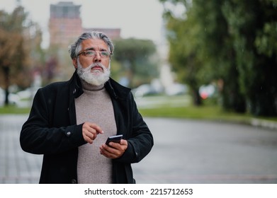 Portrait Of A Confident Mature Businessman Walking Outside While Going To A Meeting And Using A Smartphone. Senior Bearded Man Dressed In Stylish Black Coat Typing On A Mobile Phone. Copy Space.