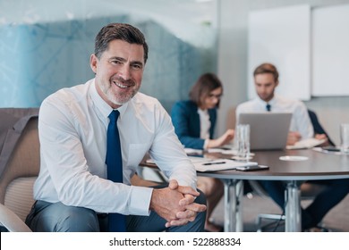 Portrait of a confident mature businessman wearing a shirt and tie sitting at a table in an office with colleagues working in the background