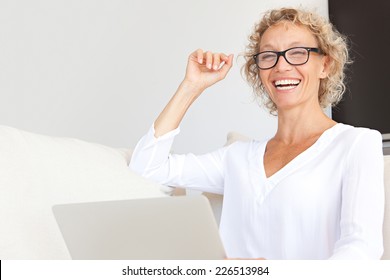 Portrait Of A Confident Mature Professional Woman Sitting On A White Sofa At Home Using A Laptop Computer To Work, Joyfully Laughing Indoors. Businesswoman Wearing Glasses And Working From Home.