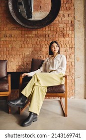 Portrait Of Confident Mid Adult Chinese Businesswoman Sitting On Retro Chair In Office With Exposed Brick Wall Interior