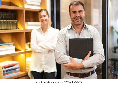 Portrait of confident mid adult executive with secretary standing behind. Caucasian bearded man holding folder, looking at camera and smiling in his office. Success concept