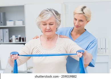 Portrait of confident nurse assisting senior woman in exercising with resistance band in clinic