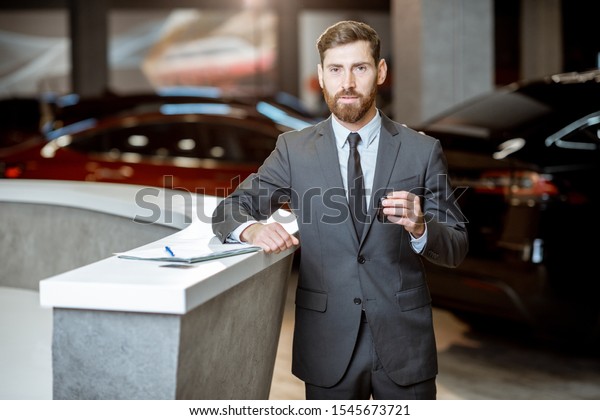 Portrait of a confident sales manager in the\
suit standing with car key in the showroom with electric cars on\
the background