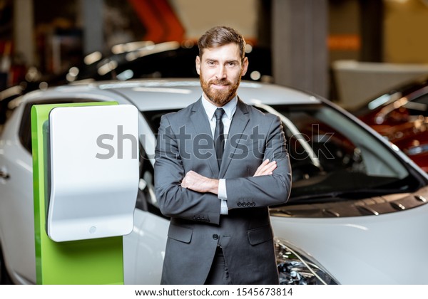 Portrait\
of a confident sales manager in the suit near the car charging\
station, selling electric cars in the\
showroom