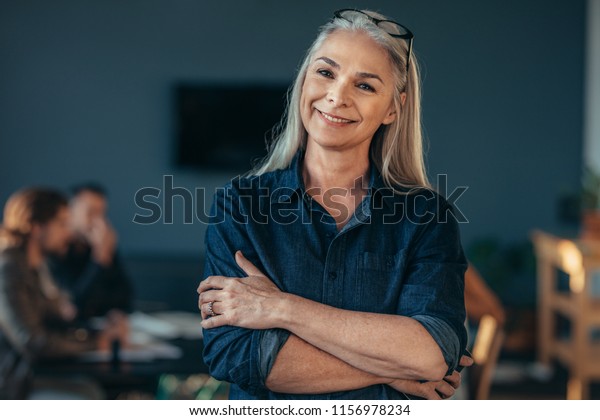 Portrait of confident senior business woman\
standing in office with her arms crossed. Mature female in office\
with colleagues discussing work at the\
back.
