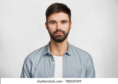 Portrait of confident serious male model has black beard and mustache, dressed in casual denim shirt, looks seriously into camera, isolated over white background. Hipster guy models in white studio - Powered by Shutterstock