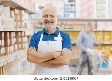 Portrait of a confident smiling grocery clerk at the supermarket, he is posing with arms crossed, store shelves in the background - Powered by Shutterstock