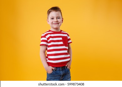 Portrait Of A Confident Smiling Little Kid Standing With Hands In Pockets Isolated Over Orange Background