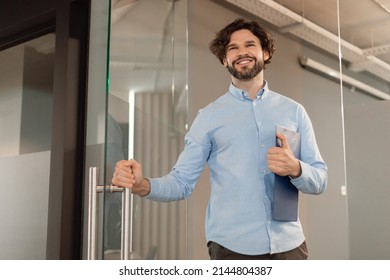 Portrait Of Confident Smiling Middle Aged Business Man In Shirt Opening Glass Door Walking In Modern Office And Smiling, Holding Clipboard And Handle, Free Copy Space, Low Angle View