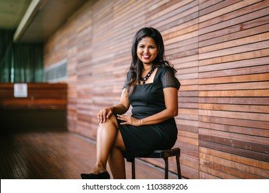 Portrait Of A Confident, Successful And Attractive Indian Asian Business Woman Sitting On A Chair In A Corridor Of Her Office In The Day. She Is Smiling For Her Professional Head Shot For Linkedin.