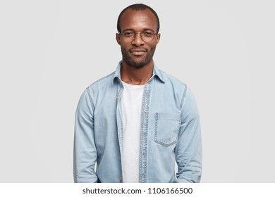 Portrait of confident successful businessman dressed in casual denim shirt, has dark skin, looks self assured, wears big glasses, stands against white background, waits for partners to have meeting