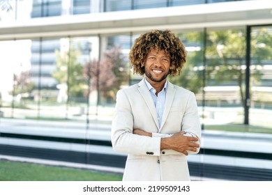 Portrait Of A Confident Successful Elegant Curly Brazilian Or Latino Man, Company Executive, Stand Outdoors Against The Backdrop Of A Business Center With Crossed Arms, Looking At Camera, Smiling