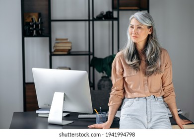 Portrait Of A Confident Successful Mature Gray-haired Asian Woman, Business Lady, Leader Or Coach Standing In Modern Office Near Work Desk, Wearing A Stylish Clothes, Looking The Side, Smiles