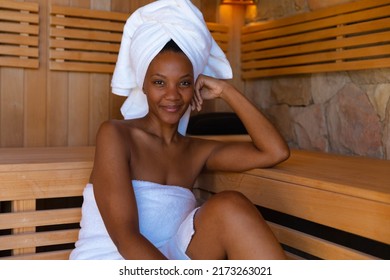 Portrait of confident young african american woman wrapped in towels sitting in sauna. Unaltered, spa, home, relaxation, pampering, detox, body care and wellbeing concept.