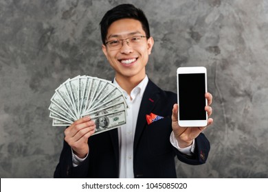 Portrait Of A Confident Young Asian Man Dressed In Suit Holding Bunch Of Money Banknotes While Showing Blank Screen Mobile Phone Over Gray Background