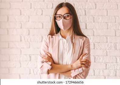 Portrait selbstbewusste junge Unternehmerin in Schutzmaske, Anzug und Brille halten die Arme gekreuzt. Schöne professionelle Geschäftsfrau in Gläsern mit rosa medizinischer Gesichtsmaske. Girl-Chef