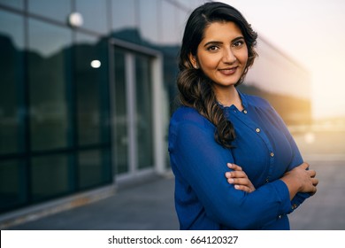 Portrait of a confident young Indian businesswoman standing with her arms crossed outside on an office building balcony overlooking the city at dusk