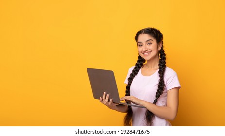 Portrait Of Confident Young Indian Woman Holding And Using Laptop Computer Isolated Over Bright Yellow Orange Studio Background With Free Copy Space. Smiling Lady Standing With Pc, Looking At Camera