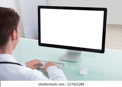 Portrait Of Confident Young Male Doctor Working On Computer At Desk In Hospital